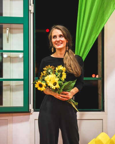 Eine Frau steht mit gelben Blumen in der Hand vor einem geöffneten Fenster und lächelt